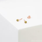 The Birthstone Studs are small, round gold earrings with green gemstones, shown on a white surface—ideal for everyday wear.