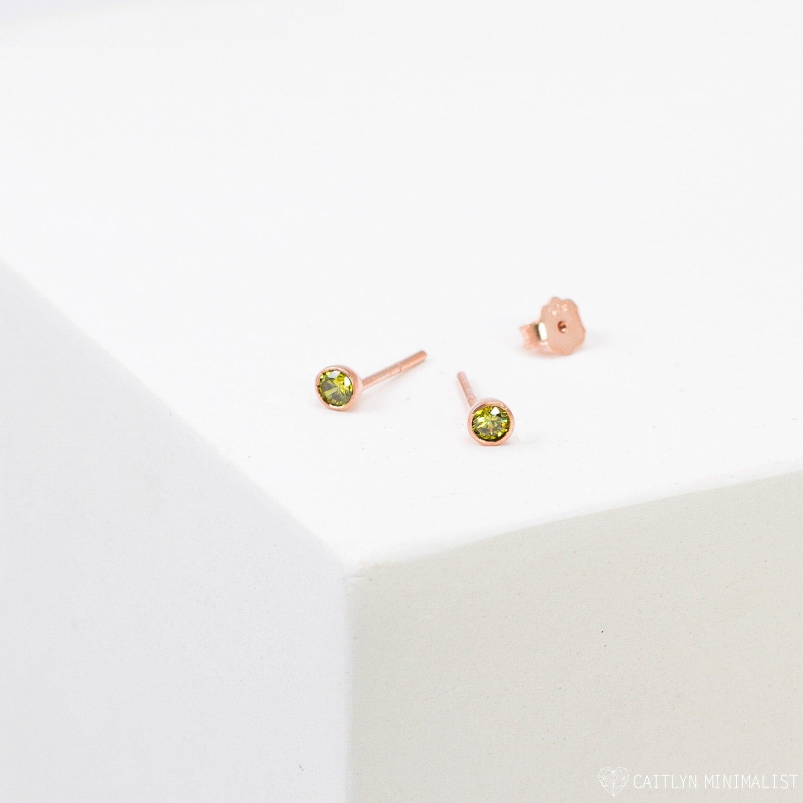 The Birthstone Studs are small, round gold earrings with green gemstones, shown on a white surface—ideal for everyday wear.