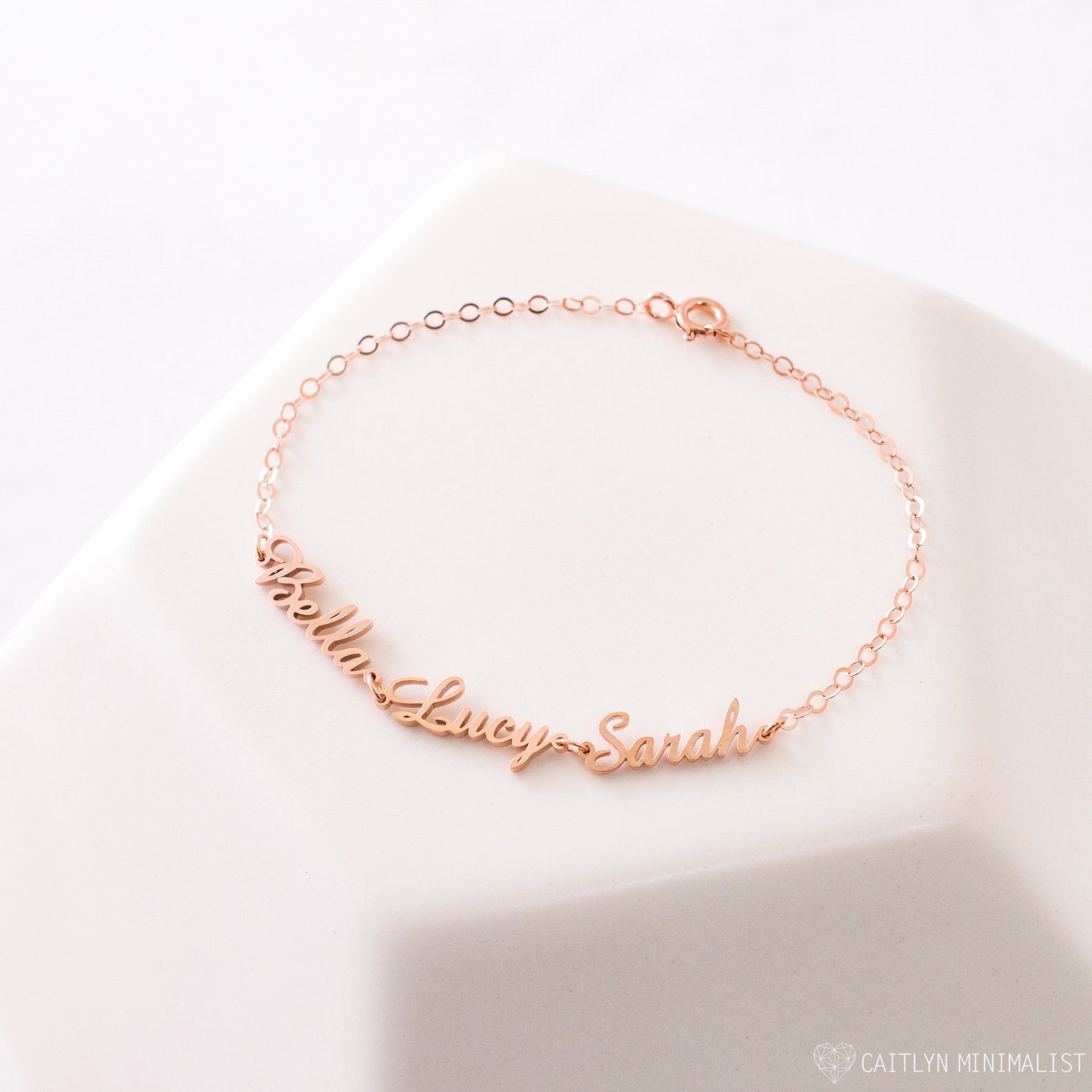The Classic Multiple Name Bracelet in rose gold features "Bella," "Lucy," and "Sarah" in elegant cursive on a dainty chain—a perfect personalized jewelry piece.