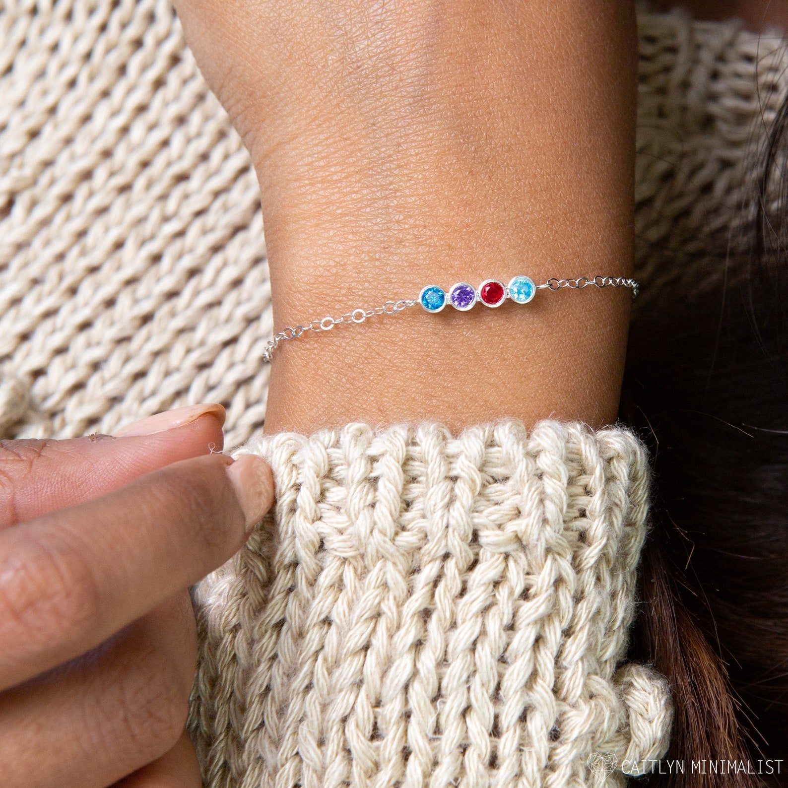 A hand wearing the Multiple Birthstone Bracelet, crafted from sterling silver and featuring four colorful personalized birthstones, rests on a chunky knit sweater.