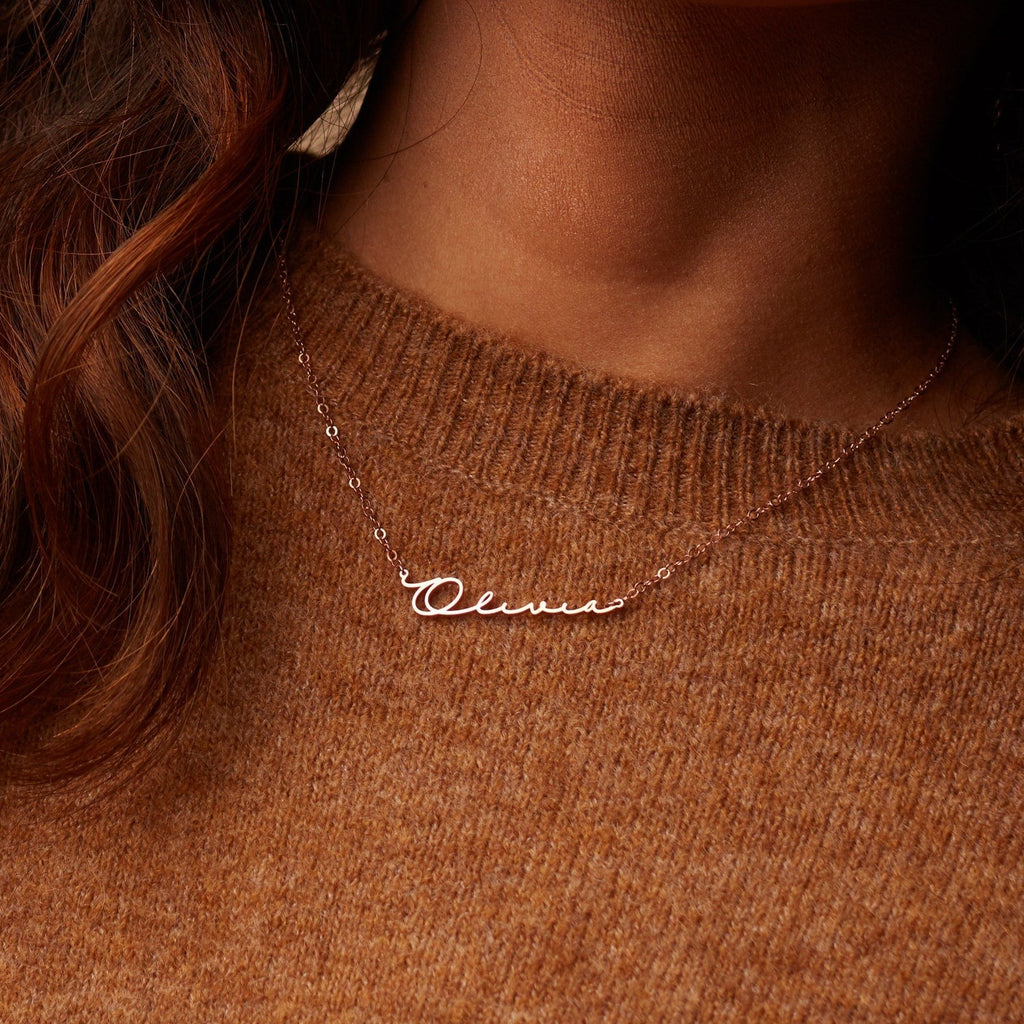 A woman wears the Minimal Name Necklace featuring "Olivia" in cursive, paired with a brown knit sweater for a chic, understated style.