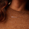 A woman wears the Minimal Name Necklace featuring "Olivia" in cursive, paired with a brown knit sweater for a chic, understated style.