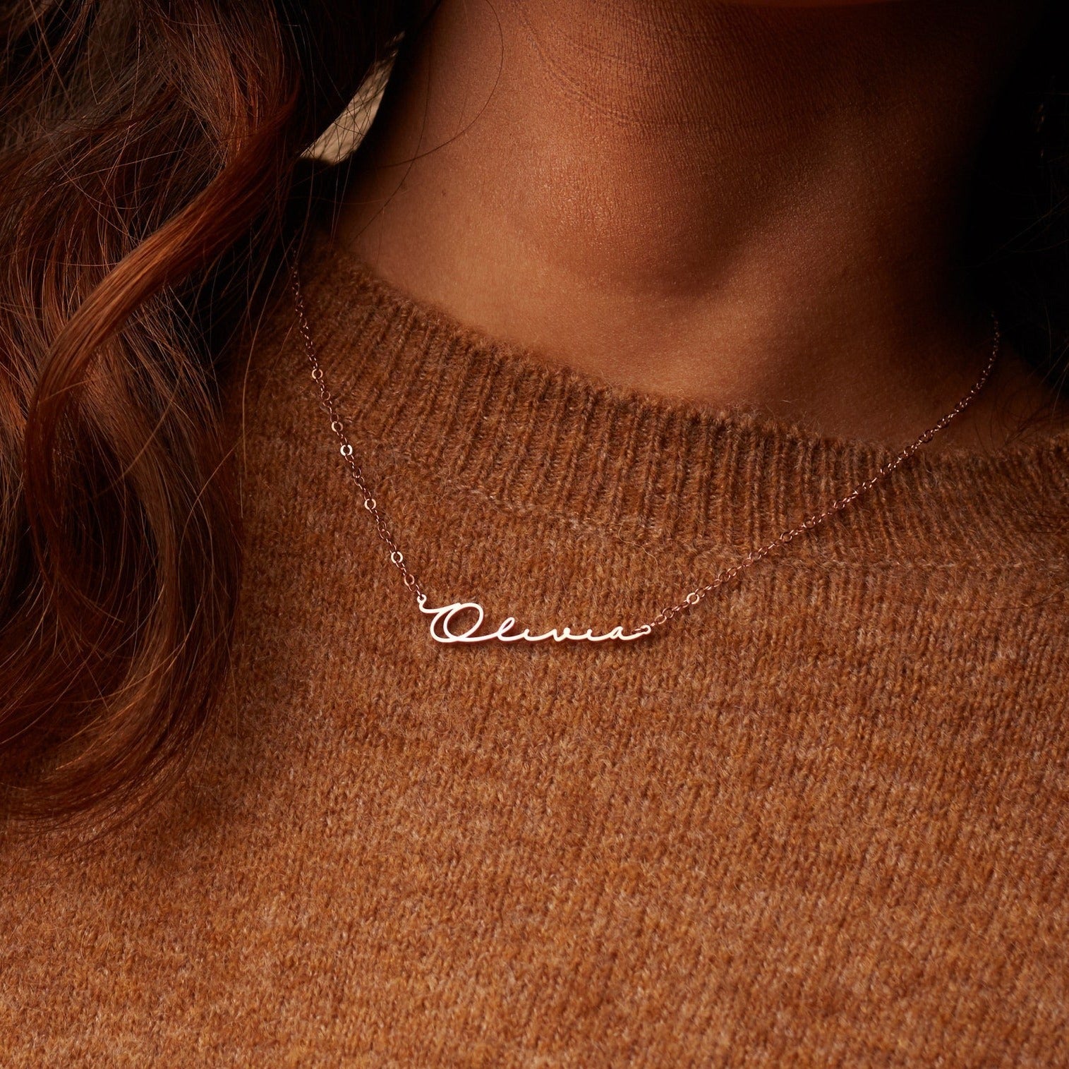 A woman wears the Minimal Name Necklace featuring "Olivia" in cursive, paired with a brown knit sweater for a chic, understated style.