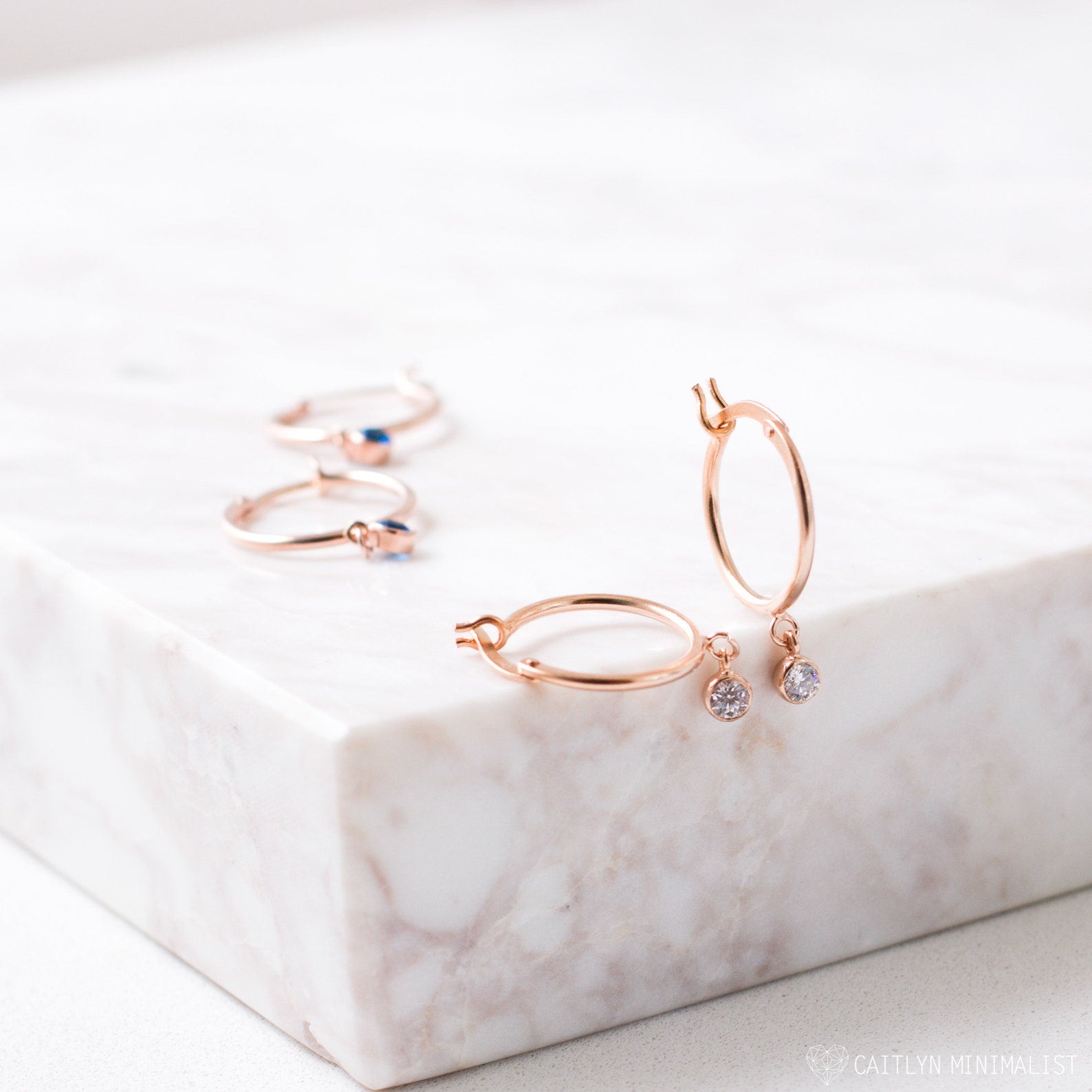 Birthstone Huggies—rose gold dainty huggie hoop earrings with dangling crystals and matching rings—displayed on a white marble surface.