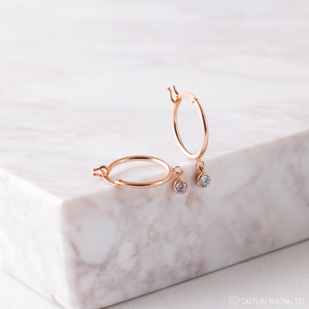 Birthstone Huggies—dainty huggie hoop earrings with small dangling clear gems—are displayed on a light marble surface and add a subtle sparkle to any look.