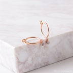 Birthstone Huggies—dainty huggie hoop earrings with small dangling clear gems—are displayed on a light marble surface and add a subtle sparkle to any look.