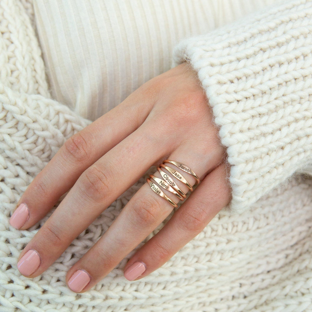 A hand with pink nails wears stackable Arthur Skinny Name Rings engraved with names, resting on a cream knit sweater.