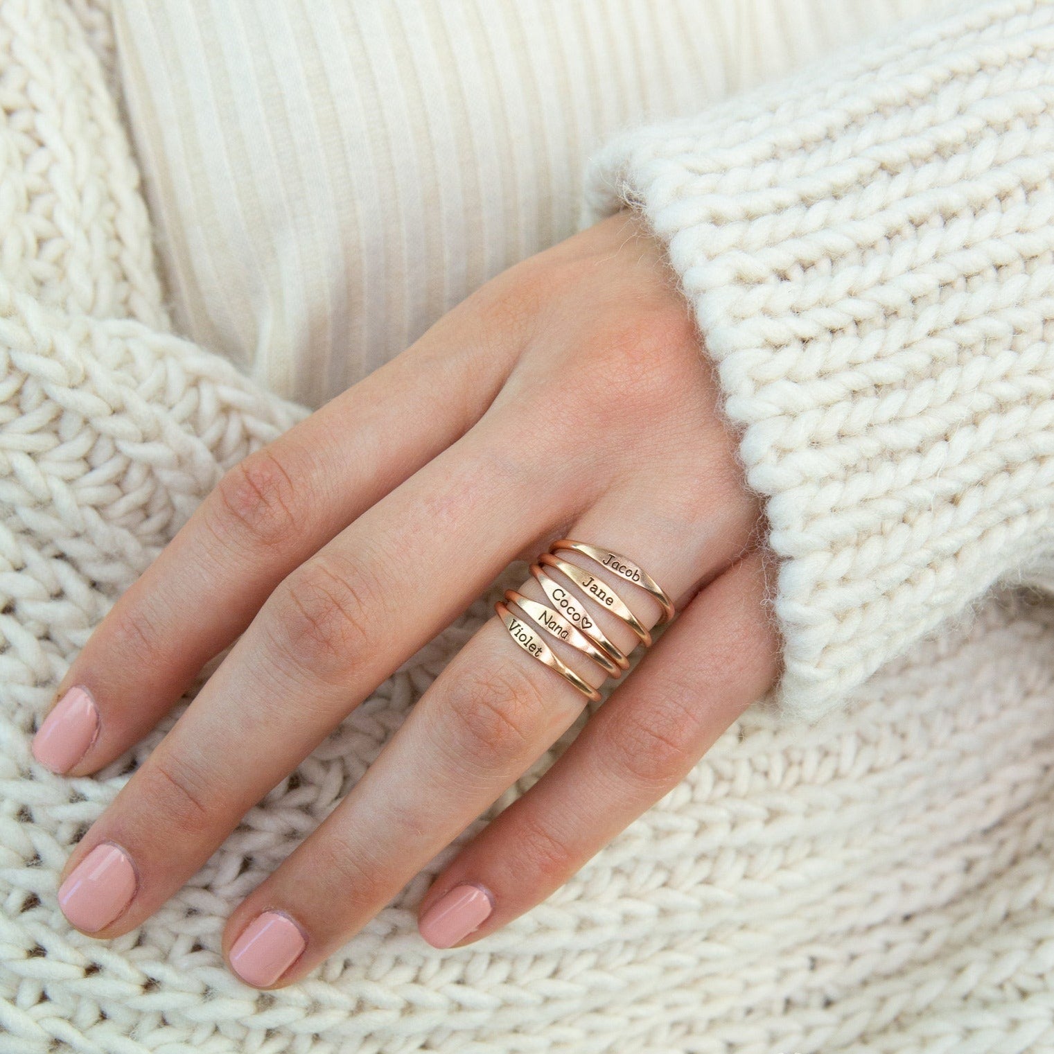 A hand with pink nails wears stackable Arthur Skinny Name Rings engraved with names, resting on a cream knit sweater.