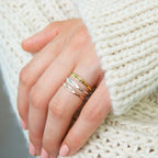 A hand with pink nails wears the Arthur Skinny Name Ring and two engraved bands, resting on a chunky knit cream sweater.