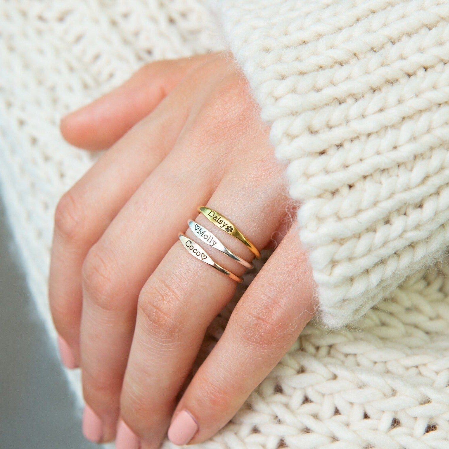 A hand with pink nails wears the Arthur Skinny Name Ring and two engraved bands, resting on a chunky knit cream sweater.