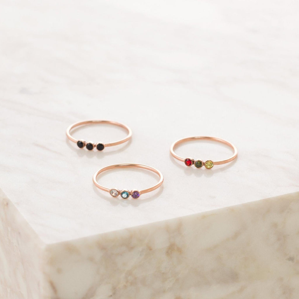 Three delicate gold rings, each a Tiny Triple Birthstone Ring with three small colored stones, displayed on light marble. Perfect for wearing solo or stacking with other thin bands.