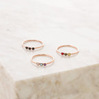 Three delicate gold rings, each a Tiny Triple Birthstone Ring with three small colored stones, displayed on light marble. Perfect for wearing solo or stacking with other thin bands.