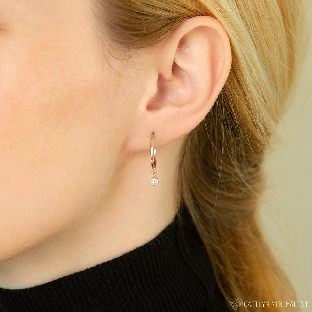Close-up of a woman's ear wearing Birthstone Huggies, featuring a delicate gold hoop with a dangling round diamond—perfect for those who love personalized earrings.