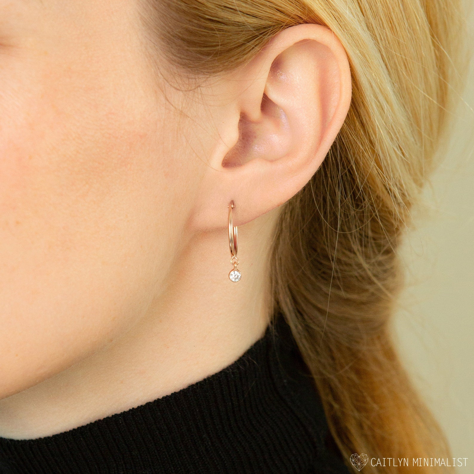 Close-up of a woman's ear wearing Birthstone Huggies, featuring a delicate gold hoop with a dangling round diamond—perfect for those who love personalized earrings.