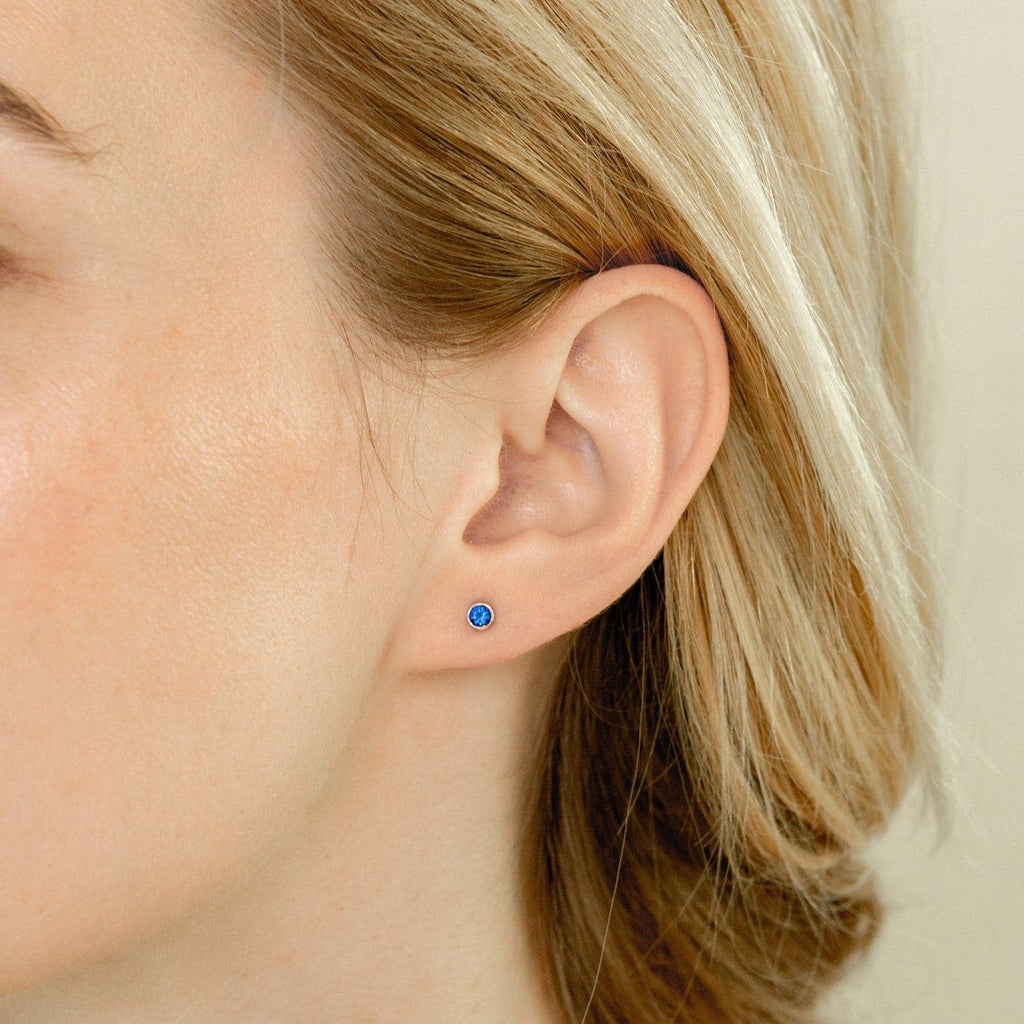 A woman with blonde hair tucked behind her ear showcases the Birthstone Studs—a delicate blue birthstone earring, perfect for everyday wear.