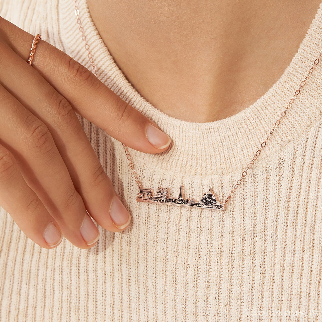 A hand adjusts the City Skyline Necklace, featuring a cityscape pendant, worn over a cream ribbed sweater—an ideal personalized gift.