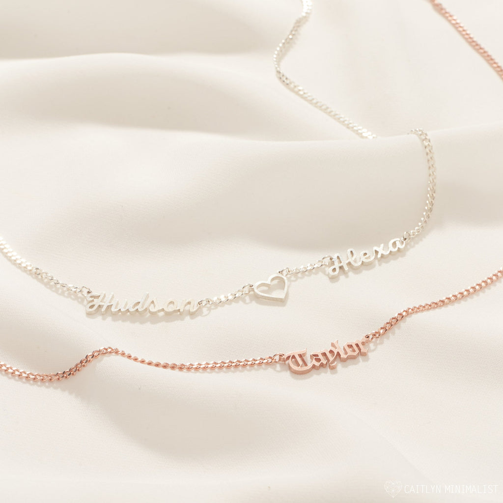 Two Old English Name Necklaces in Curb Chain on a cream background—one silver, one rose gold with a heart detail.
