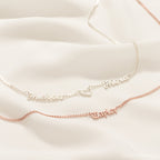 Two Old English Name Necklaces in Curb Chain on a cream background—one silver, one rose gold with a heart detail.