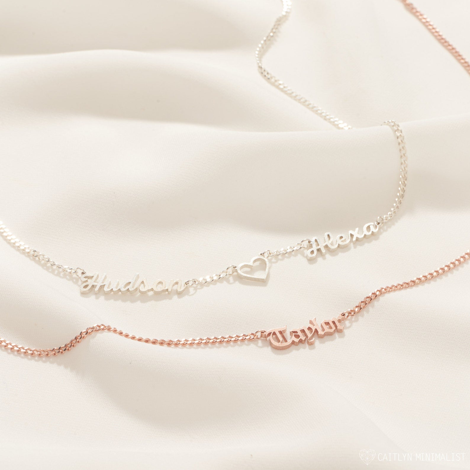 Two Old English Name Necklaces in Curb Chain on a cream background—one silver, one rose gold with a heart detail.