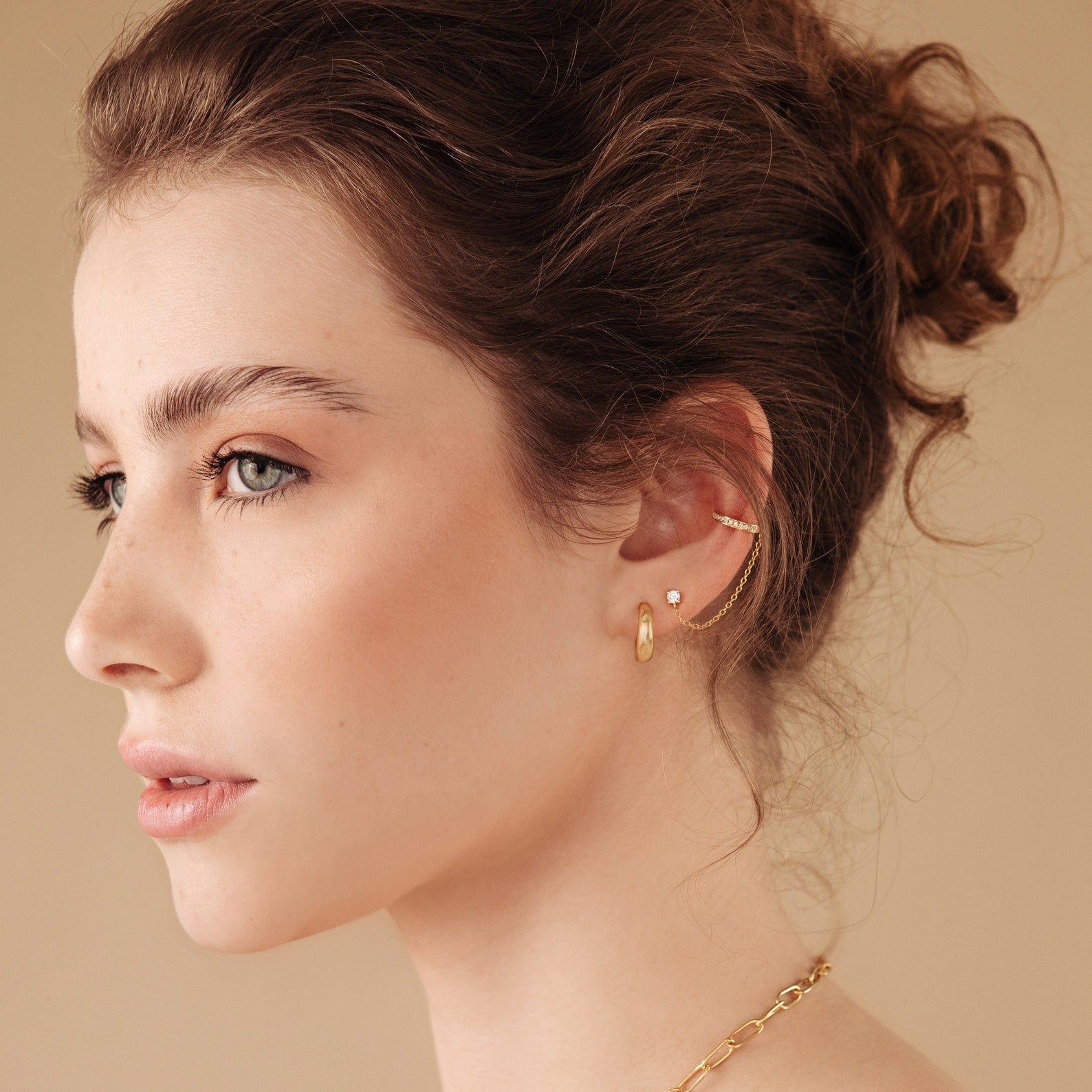 A woman with wavy brown hair models Pave Chain Ear Cuffs in Rose Gold against a beige background.