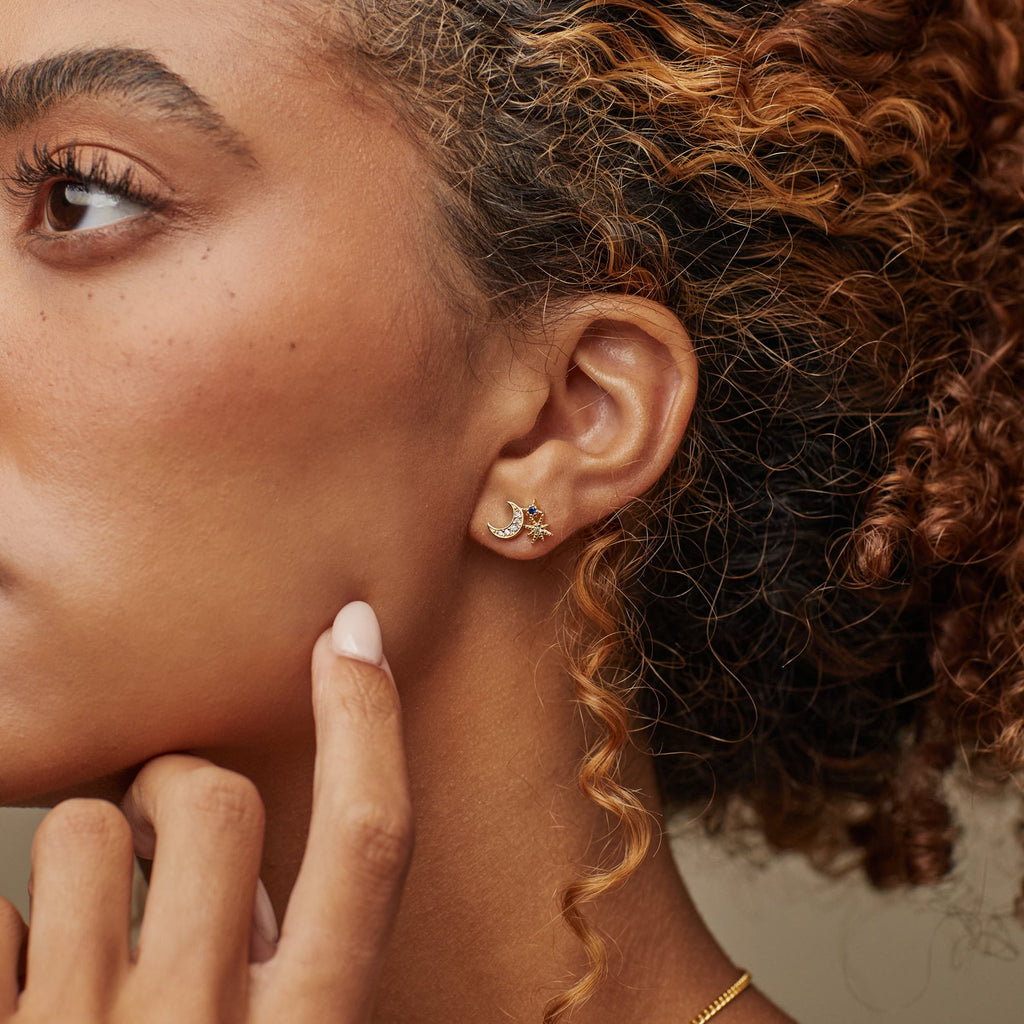 Woman with curly hair wearing a gold crescent moon earring accented with a star, touching her face gently—an elegant style that pairs perfectly with Aeon Studs or other constellation-inspired earrings.