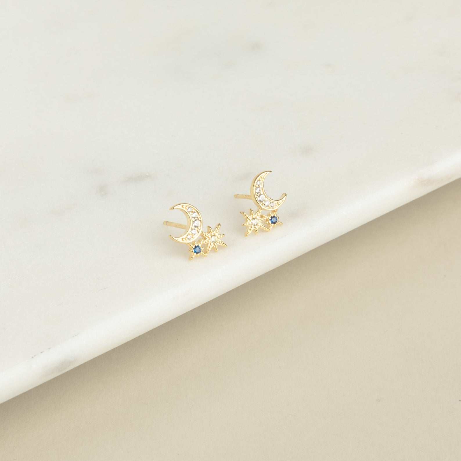 Aeon Studs: Gold constellation earrings shaped like crescent moons and stars, accented with blue stones that evoke sapphire, shown on a marble surface.