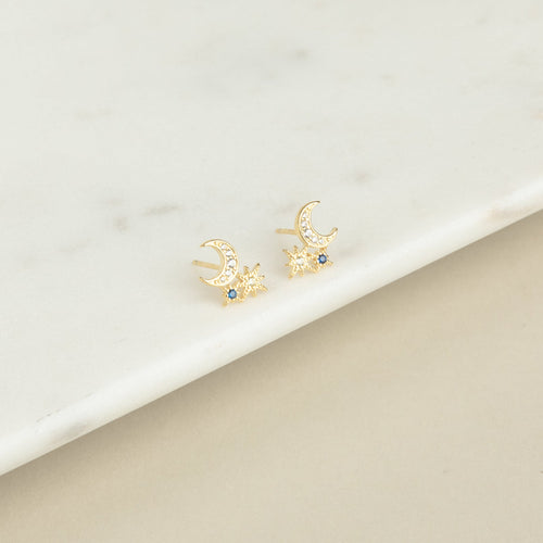 Aeon Studs: Gold constellation earrings shaped like crescent moons and stars, accented with blue stones that evoke sapphire, shown on a marble surface.