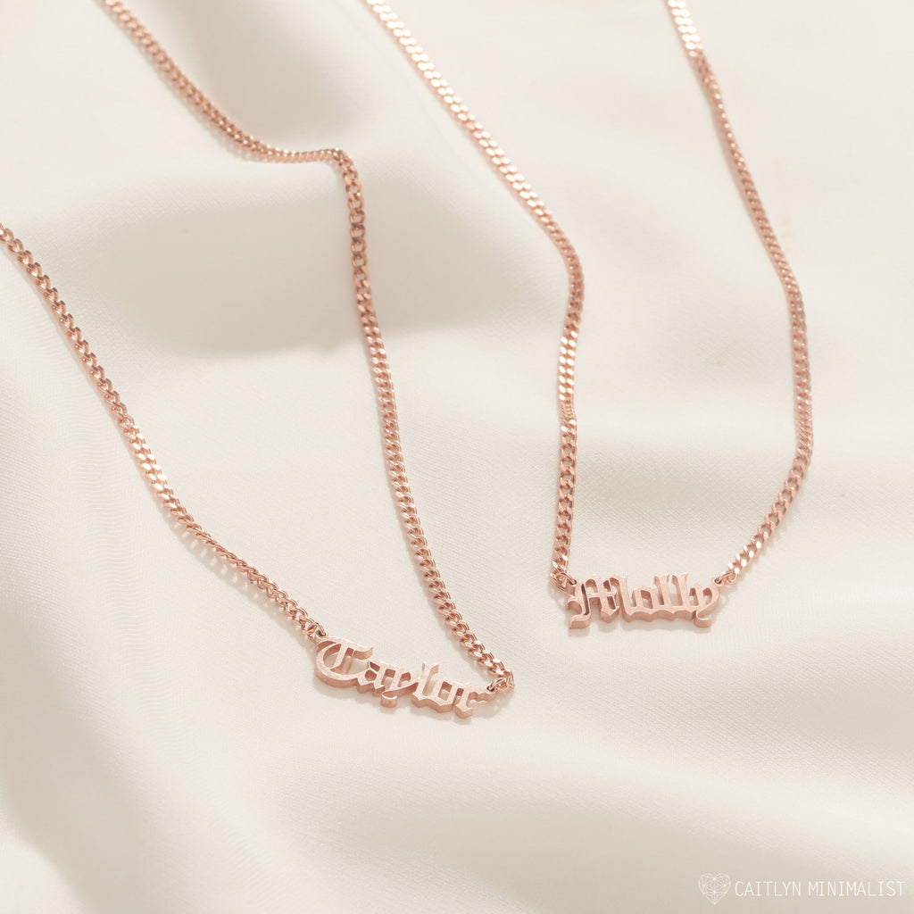 Two Old English Name Necklaces in Curb Chain, each with "Taylor" and "Milly" pendants in rose gold, rest on soft beige fabric, highlighting their personalized design and elegant curb chains.