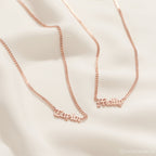 Two Old English Name Necklaces in Curb Chain, each with "Taylor" and "Milly" pendants in rose gold, rest on soft beige fabric, highlighting their personalized design and elegant curb chains.