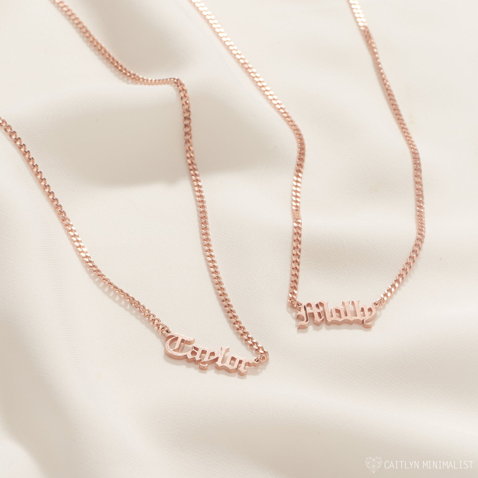 Two Old English Name Necklaces in Curb Chain, each with "Taylor" and "Milly" pendants in rose gold, rest on soft beige fabric, highlighting their personalized design and elegant curb chains.