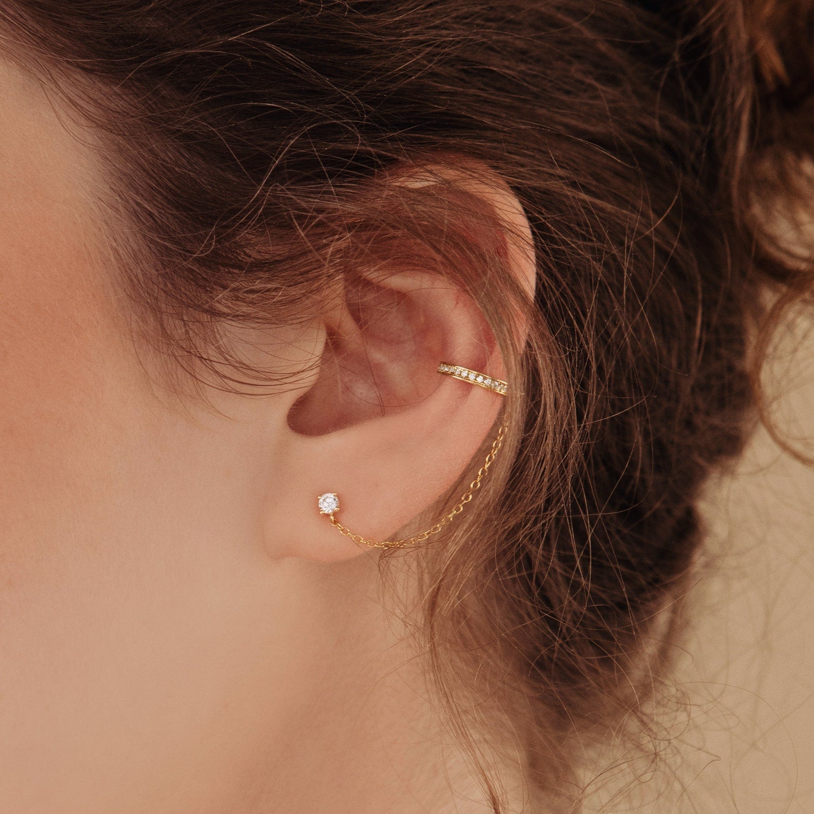 Close-up of an ear wearing Pave Chain Ear Cuffs, featuring a delicate gold chain connecting a stud to a cartilage cuff on the upper ear.