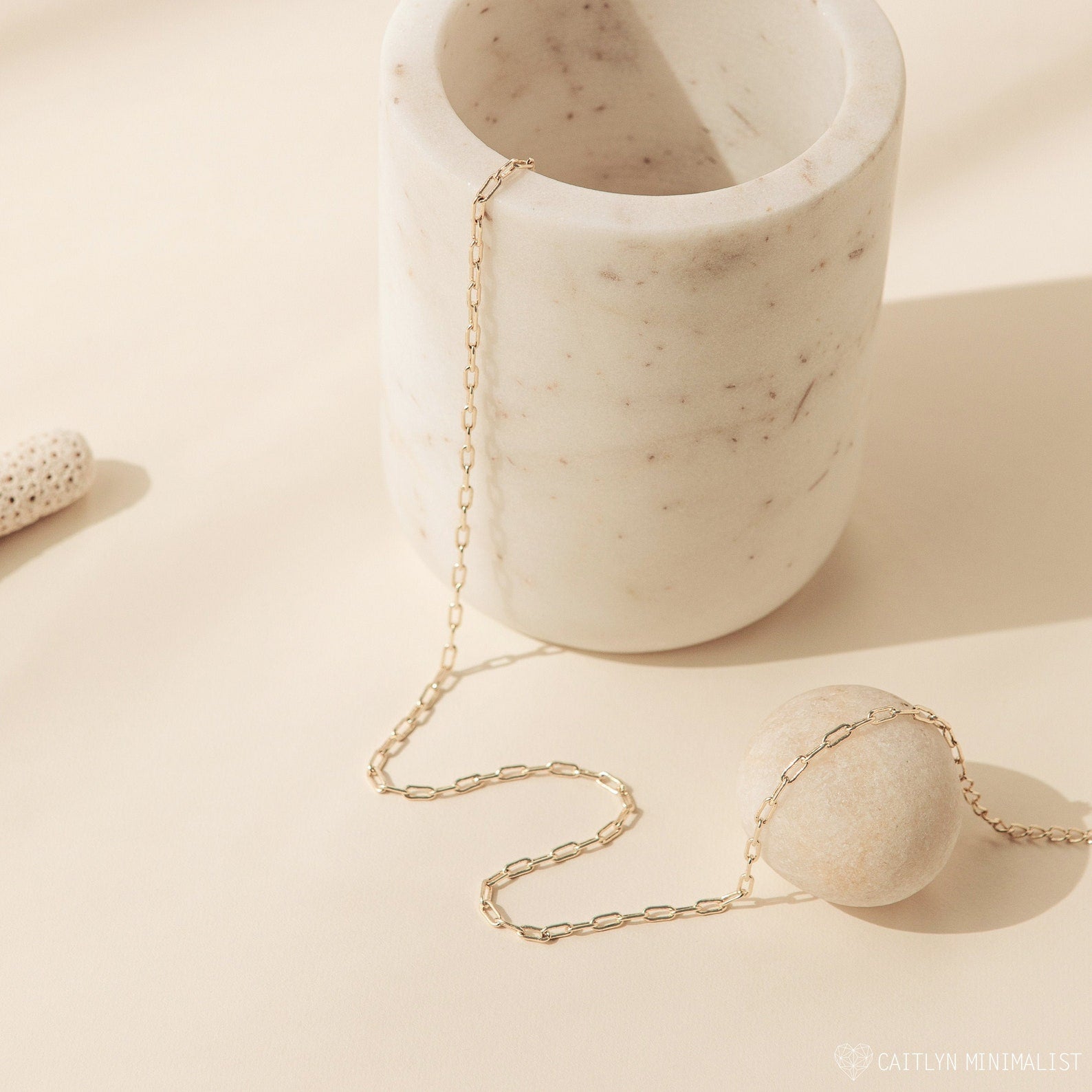 The Dainty Link Chain Necklace, a delicate gold accessory, is elegantly draped over a white marble cup and stone on a neutral background.