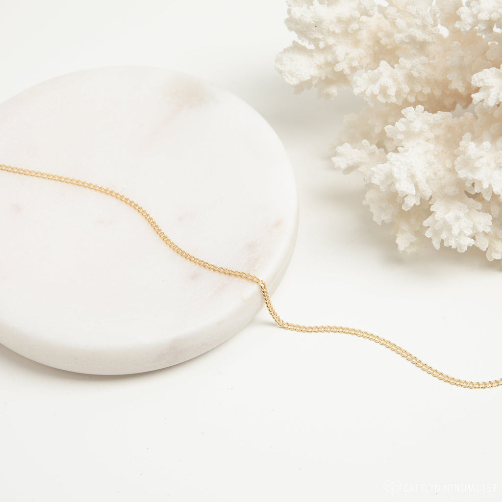 A gold curb chain necklace styled on a white marble round tray next to white coral, arranged in a flowing curve.