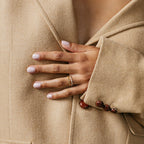 A hand with pale pink nails and gold rings, featuring the Baguette Eternity Ring, rests on a tan coat with brown buttons—a chic touch of modern luxury.