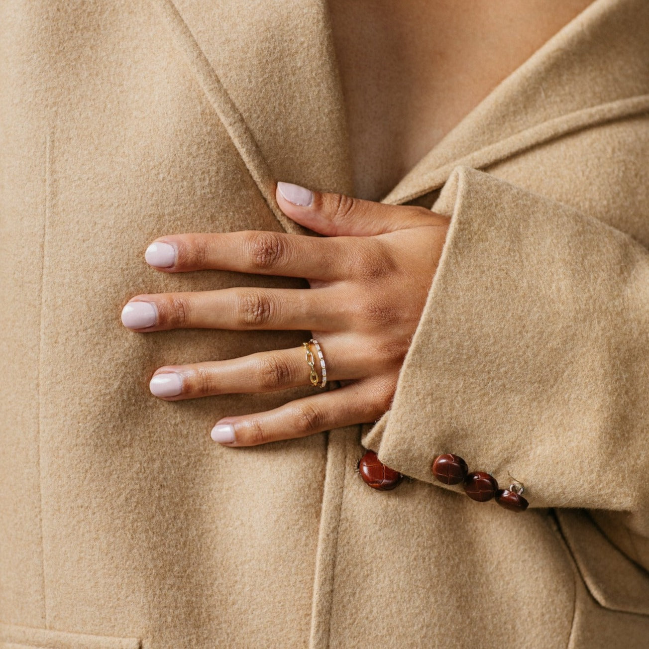 A hand with pale pink nails and gold rings, featuring the Baguette Eternity Ring, rests on a tan coat with brown buttons—a chic touch of modern luxury.