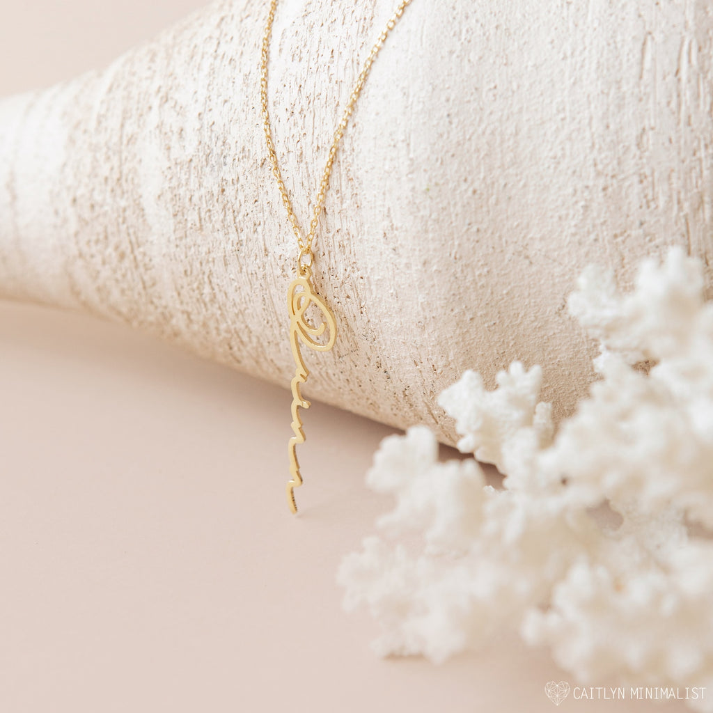 The Vertical Minimal Name Necklace is displayed on a seashell with white coral in the foreground, creating a sleek, amulet-inspired look.