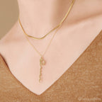 A person in a brown top wears two gold necklaces: one is a Vertical Minimal Name Necklace, and the other is a dainty chain with a wavy, amulet-like pendant.