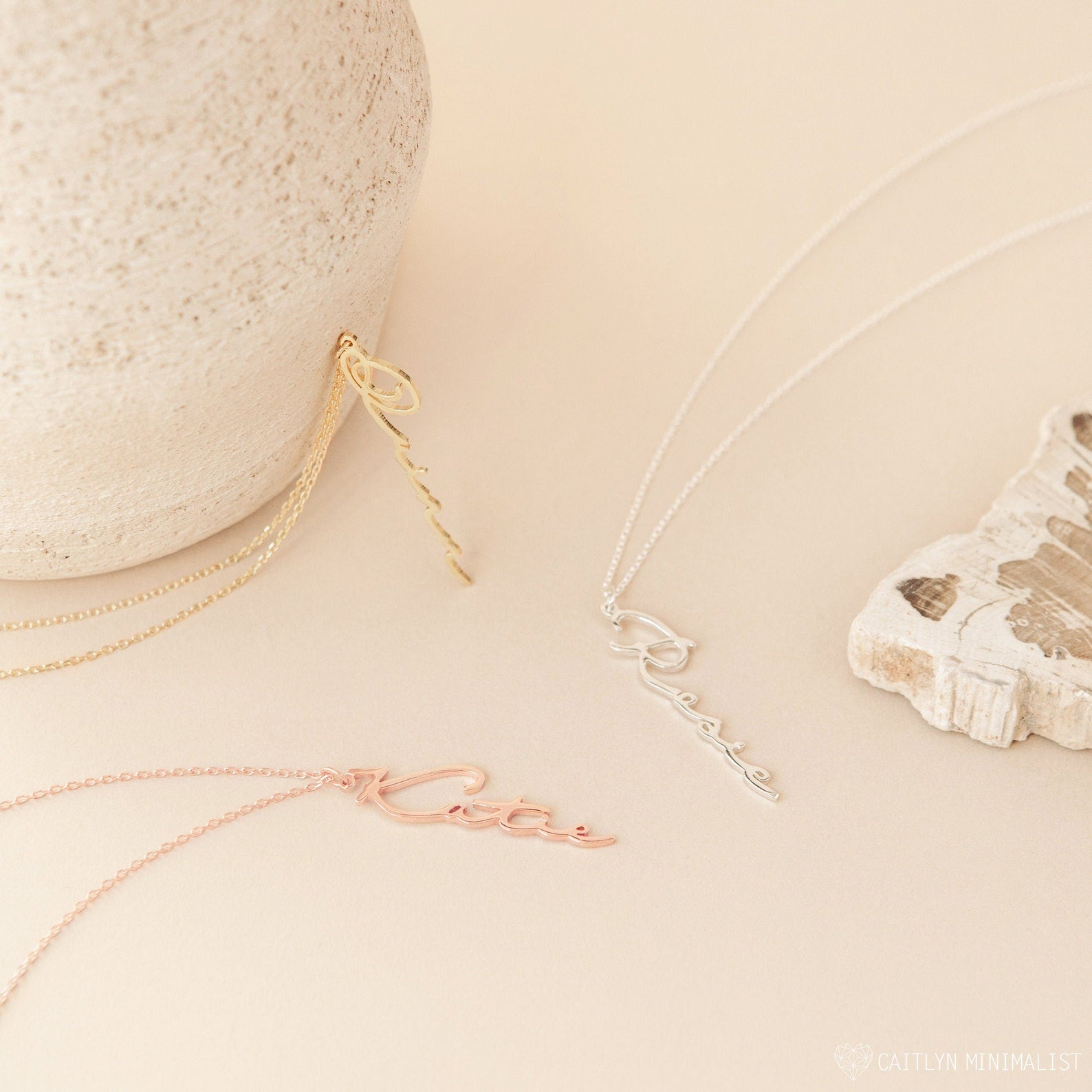 Three Vertical Minimal Name Necklaces in gold, rose gold, and silver are displayed on a beige surface with pottery, each dainty necklace serving as a chic, personalized everyday accessory.