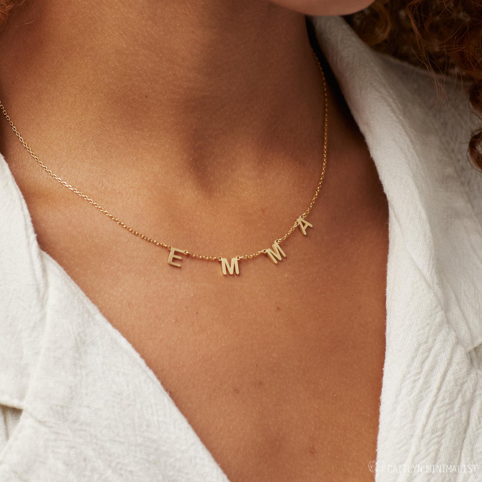 The Alice Letter Name Necklace in Rose Gold spells EMMA and is worn with a white textured shirt—making it a stylish personalized jewelry piece or thoughtful Christmas gift.
