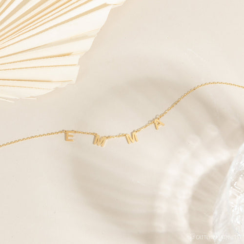 The Alice Letter Name Necklace—a gold personalized necklace with letter charms—makes a perfect Christmas gift, shown here spelling EMMA on a cream background with textured decor.