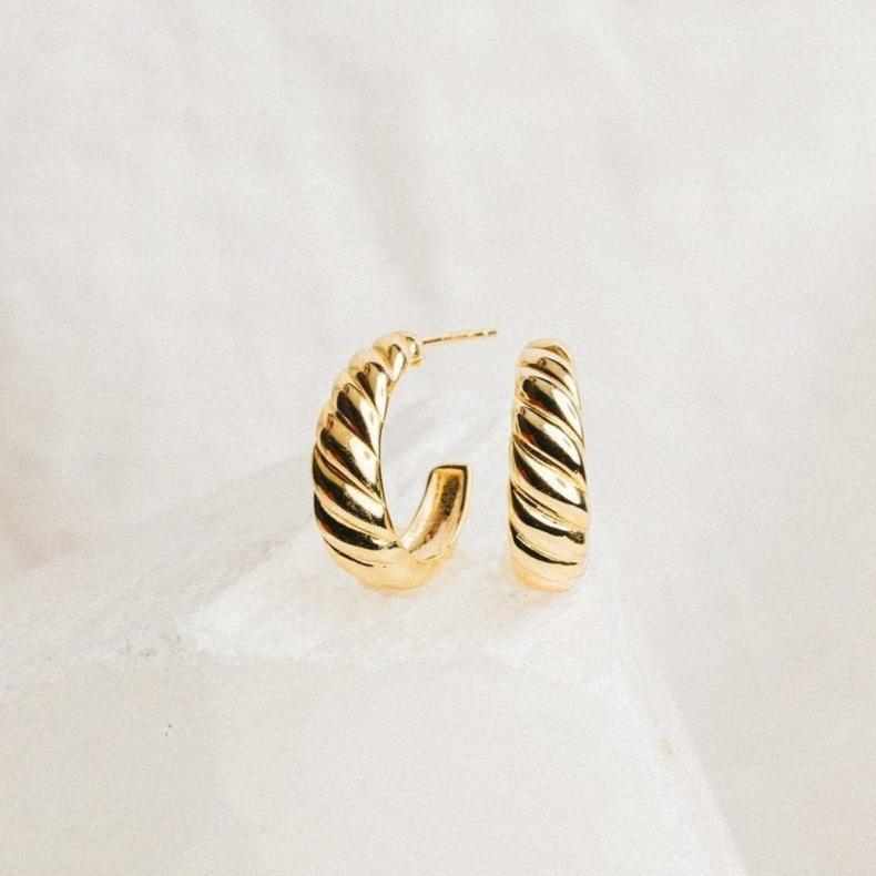 Croissant Hoops—gold twisted earrings with elegant detailing—displayed on a white textured surface.