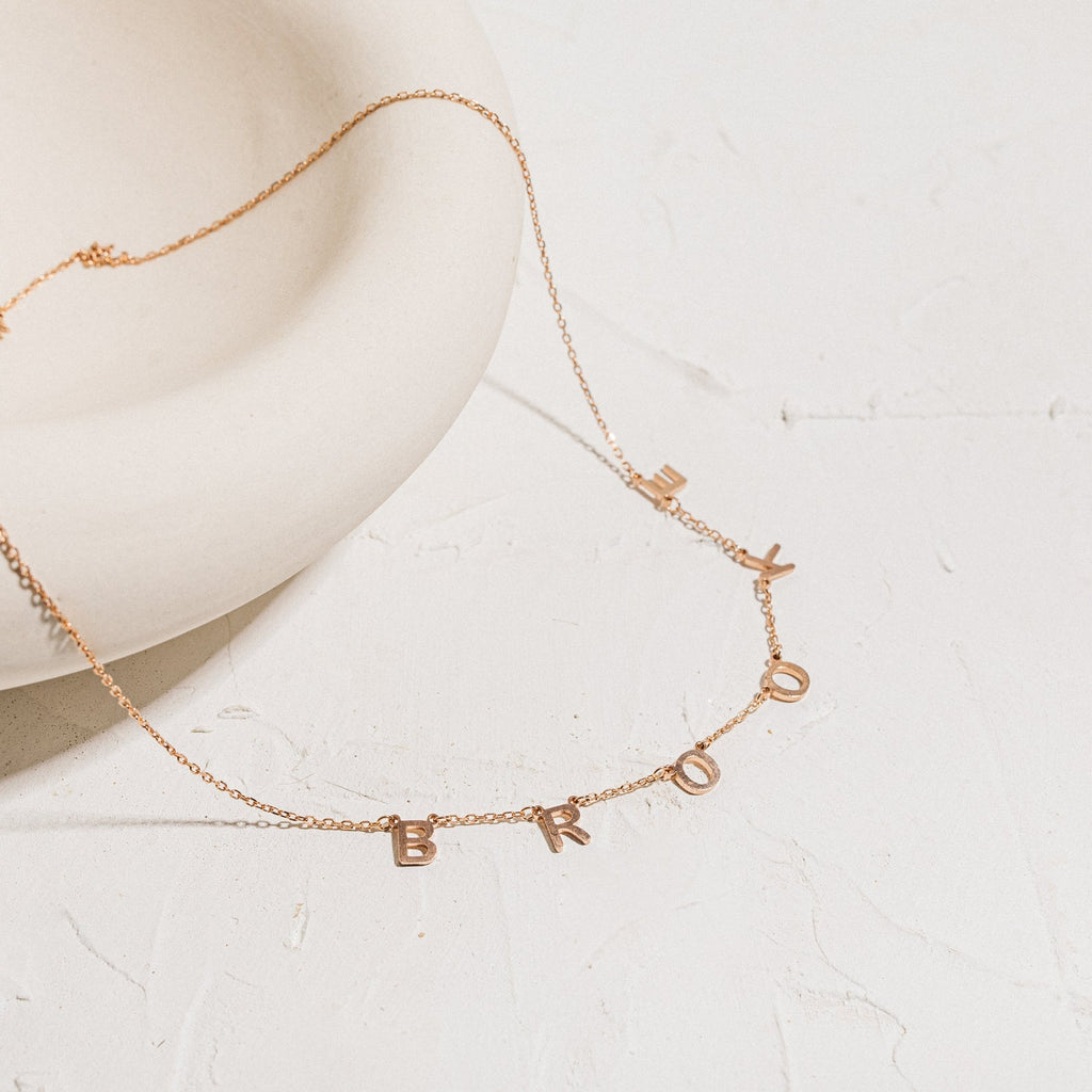 The Custom Letter Necklace in Rose Gold features dangling letter charms spelling BROOKLYN on a white textured surface—a stunning example of personalized name jewelry.