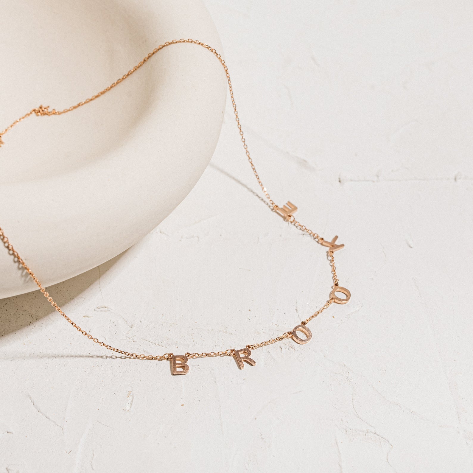 The Custom Letter Necklace in Rose Gold features dangling letter charms spelling BROOKLYN on a white textured surface—a stunning example of personalized name jewelry.
