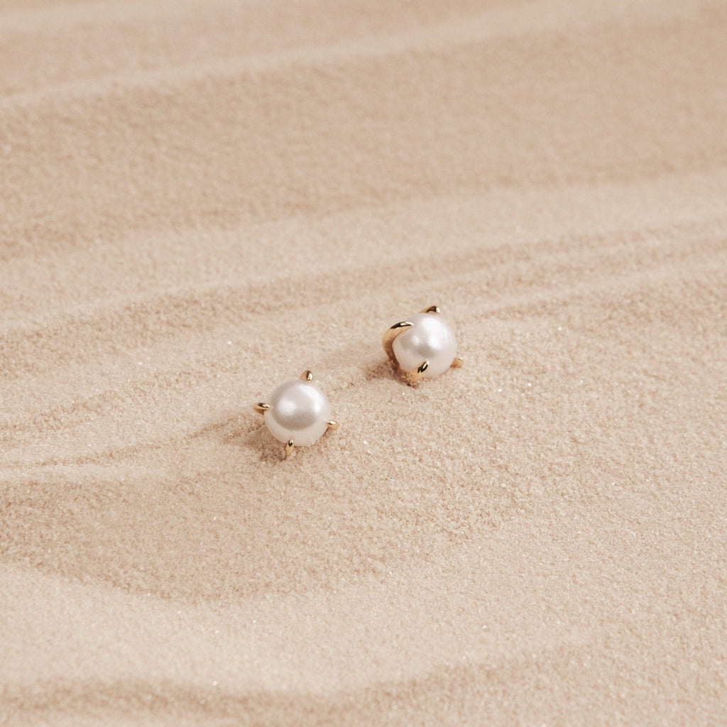 Zoe Pearl Studs with gold prongs rest on smooth, light-colored sand, reflecting the elegance of feminine jewelry and a modern style.