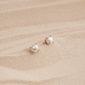 Zoe Pearl Studs with gold prongs rest on smooth, light-colored sand, reflecting the elegance of feminine jewelry and a modern style.