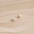 Zoe Pearl Studs with gold prongs rest on smooth, light-colored sand, reflecting the elegance of feminine jewelry and a modern style.