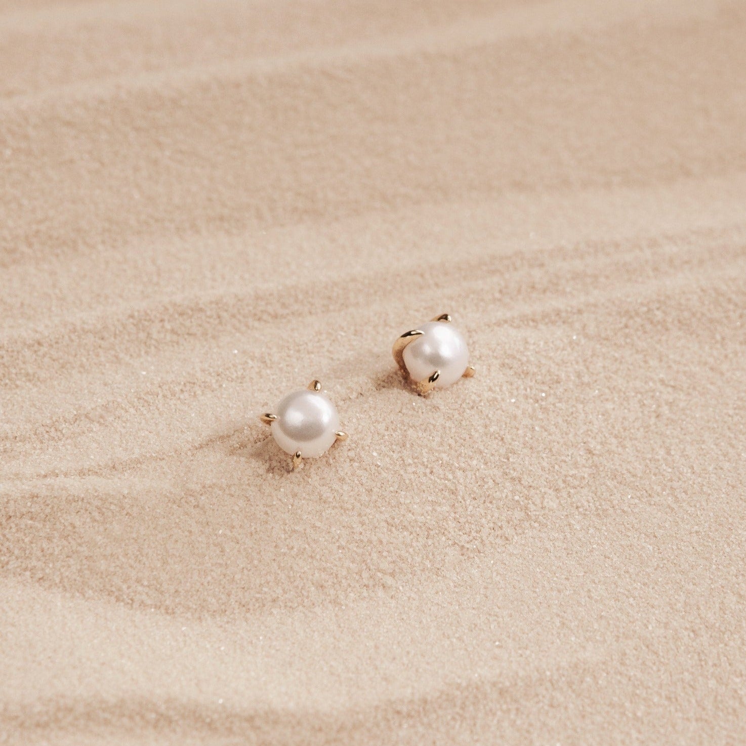 Zoe Pearl Studs with gold prongs rest on smooth, light-colored sand, reflecting the elegance of feminine jewelry and a modern style.