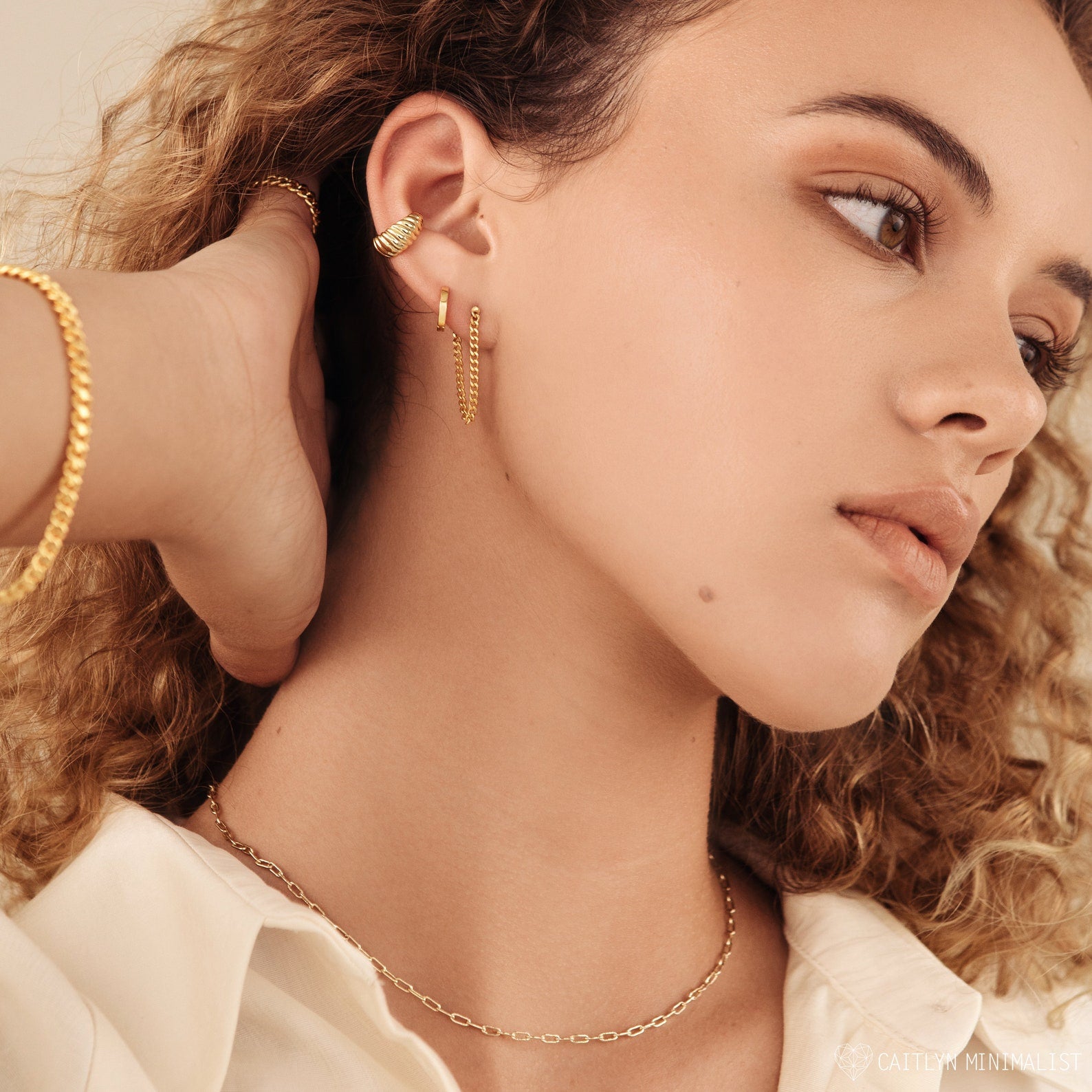 A woman with curly hair wears a white shirt, gold necklace, and versatile earrings such as the Curb Chain Studs—an ideal choice for a personalized gift.