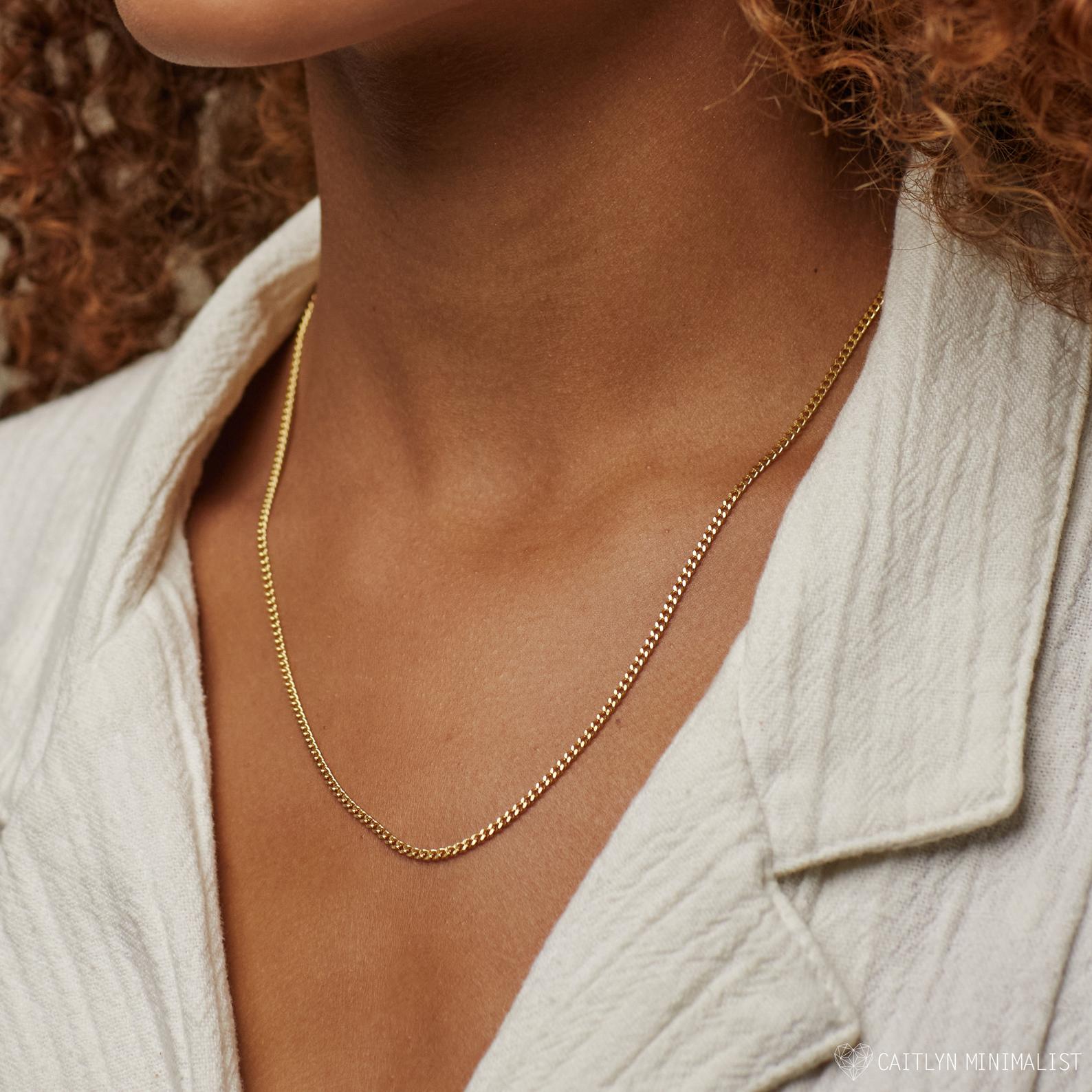 A close-up of a woman wearing a classic gold curb chain necklace against her skin. The necklace rests elegantly on her collarbone, lying flat in its distinct interlocking pattern.