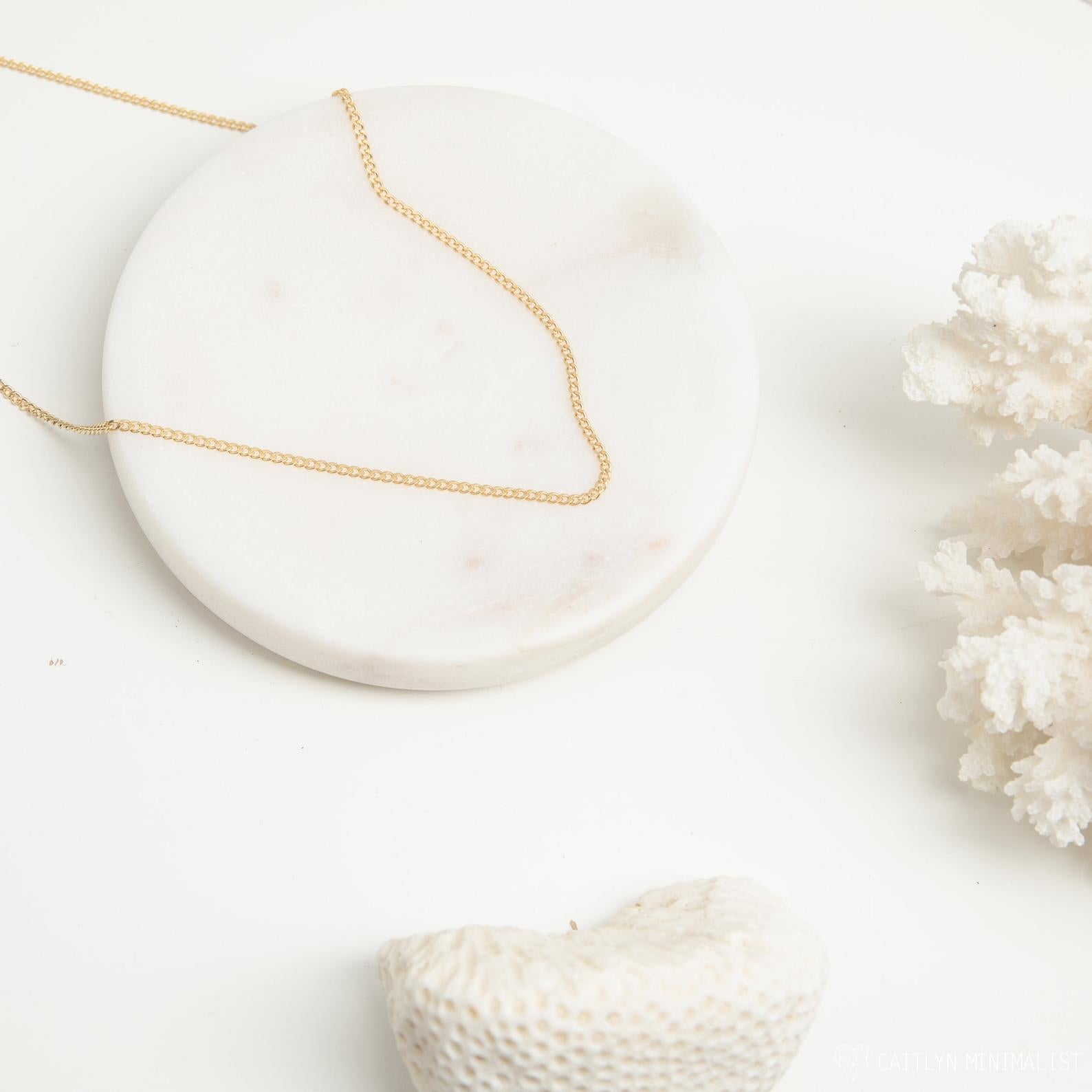 A gold curb chain necklace resting in a gentle curve on a round white marble tray with soft white coral in the background.